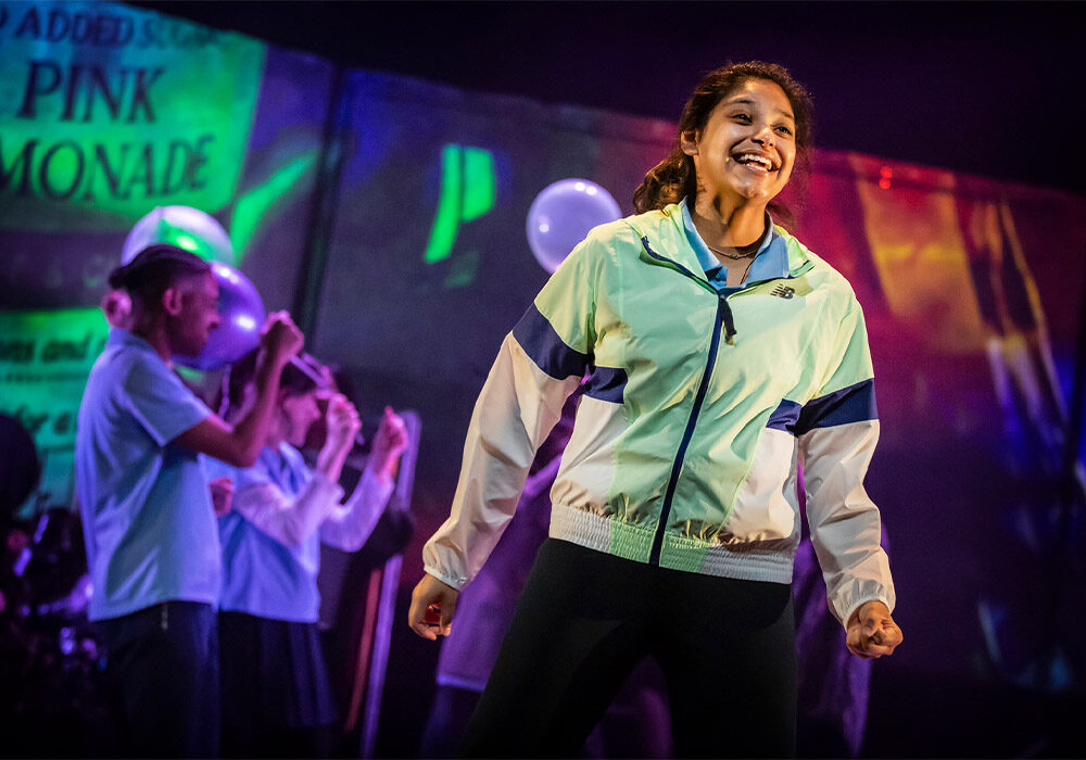 A production photo from the 2023 production of Run, Rebel. A teenage girl stands triumphantly at the front of the stage, dressed in joggers and tracksuit top. She is smiling. Behind her, out of focus,are other cast members celebrating.