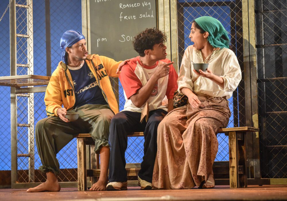Three people, two boys and one girl, sat on a bench eating from enamel bowls. The boys wear a mix of sportswear whilst the girl wears a white shirt, long skirt, and turquoise headscarf. The boy in the middle points accusingly at the girl and she is reacting in shock.