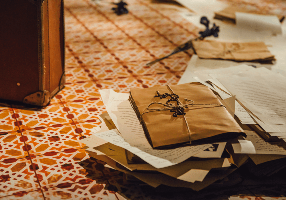 A tiled floor decorated in North African patterns with paperwork and packages stacked up. One package is tied with string and a rosary is laid across it.