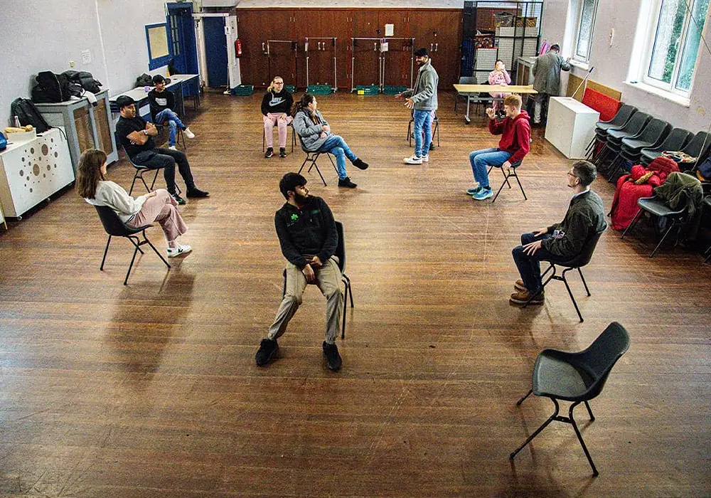 A high-angle photo of a workshop with young people. They are seated in a circle, in a hall with wooden flooring.